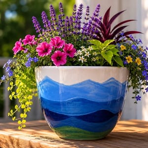 May include: A ceramic planter featuring a hand-painted landscape of blue mountain ranges and a green base. The pot is filled with a vibrant arrangement of purple lavender, bright pink petunias, small white flowers, yellow blossoms, and trailing green foliage.