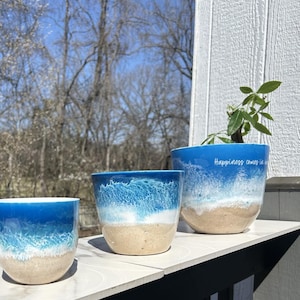 May include: Three blue and white ceramic planters with a beach scene design. The largest planter has the text "Happiness comes in waves" written on it.