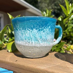 May include: A ceramic mug with a coastal design. The top half of the mug is a vibrant turquoise, transitioning to white, resembling ocean waves. The bottom half is a speckled, sandy color, creating a beach-like aesthetic. The mug has a curved handle.