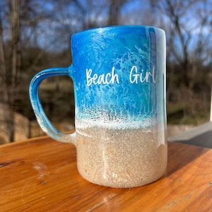 May include: A ceramic mug with a beach design. The top half is blue, like ocean waves, fading to a sandy base. The words "Beach Girl" are written in white script. The mug has a curved handle.