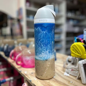 May include: A white and blue water-themed reusable water bottle with a white lid and the word "owa'o" printed on the lid. The bottle has a white inscription that says "Gabby".