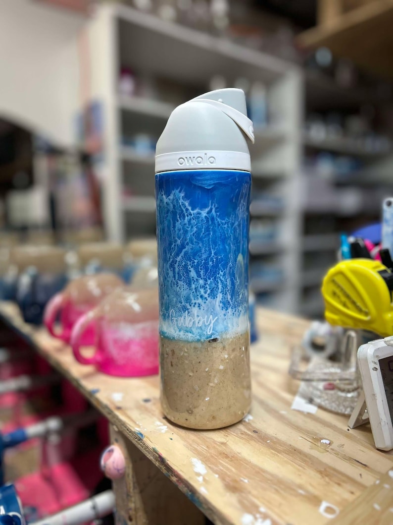 May include: A white and blue water-themed reusable water bottle with a white lid and the word "owa'o" printed on the lid. The bottle has a white inscription that says "Gabby".