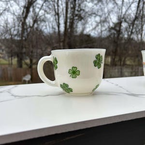 May include: A white ceramic mug with a green shamrock pattern. The mug has a handle and is sitting on a white marble surface.