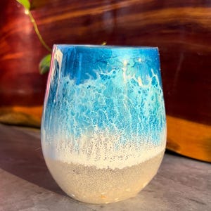 May include: A stemless glass with a beach-themed design. The top half is a vibrant blue, transitioning to white and then a sandy beige at the base. The glass has a smooth, glossy finish, evoking a coastal aesthetic.