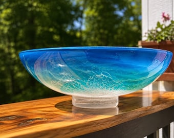 May include: A large, circular glass bowl featuring a gradient of deep blue at the rim transitioning to a lighter turquoise and white at the base. The surface displays a textured, swirling pattern that mimics ocean waves, supported by a clear glass pedestal.