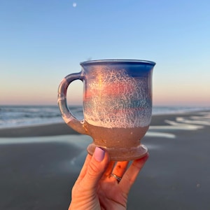 May include: A glass mug with a blue, pink, and white layered design. The mug has a handle and a rounded base. The mug is being held in a hand against a beach background.