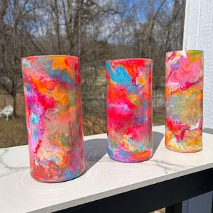 Könnte beinhalten: Drei bunte Glasbecher mit abstrakten Wirbelmustern in den Farben Rosa, Orange, Blau und Gold. Die Becher sind in einer Reihe auf einer weißen Oberfläche angeordnet.