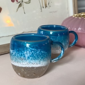 May include: Two blue and white ceramic mugs with a beach scene design. The mugs have a white rim and a white handle. The mugs are sitting on a pink surface.