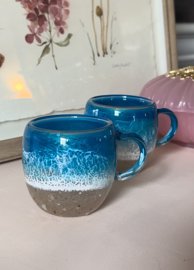 May include: Two blue and white ceramic mugs with a beach scene design. The mugs have a white rim and a white handle. The mugs are sitting on a pink surface.