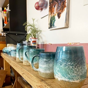 Peut inclure: Une rang&eacute;e de tasses en c&eacute;ramique avec des motifs d'ondes oc&eacute;aniques. Les tasses sont dispos&eacute;es sur une &eacute;tag&egrave;re en bois. Les tasses sont de tailles et de formes diff&eacute;rentes, mais elles ont toutes un motif d'ondes oc&eacute;aniques similaire. Les vagues sont peintes dans des tons de bleu, de vert et de blanc. Les tasses sont toutes peintes &agrave; la main et ont un aspect unique et artisanal.
