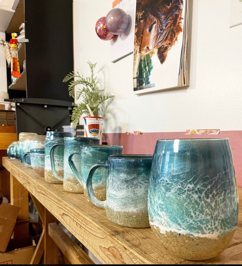 Peut inclure: Une rang&eacute;e de tasses en c&eacute;ramique avec des motifs d'ondes oc&eacute;aniques. Les tasses sont dispos&eacute;es sur une &eacute;tag&egrave;re en bois. Les tasses sont de tailles et de formes diff&eacute;rentes, mais elles ont toutes un motif d'ondes oc&eacute;aniques similaire. Les vagues sont peintes dans des tons de bleu, de vert et de blanc. Les tasses sont toutes peintes &agrave; la main et ont un aspect unique et artisanal.
