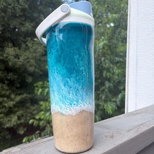May include: A water bottle with a blue and white ocean wave design. The top of the bottle is light blue, with a white handle and a light green accent. The bottom of the bottle has a tan sand design.