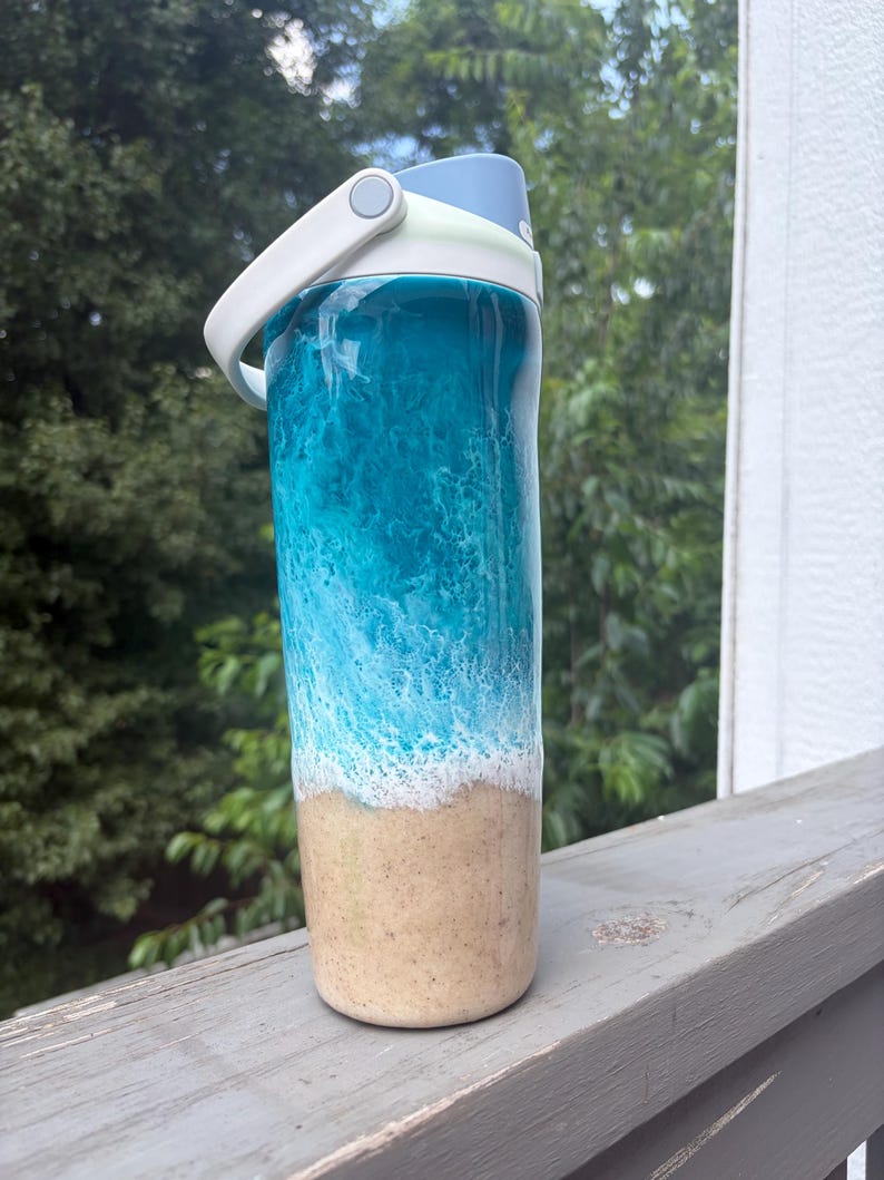 May include: A water bottle with a blue and white ocean wave design. The top of the bottle is light blue, with a white handle and a light green accent. The bottom of the bottle has a tan sand design.
