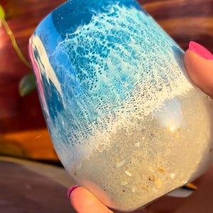 May include: A clear glass tumbler with a blue and white ocean wave design. The bottom of the tumbler is filled with white sand and small seashells.