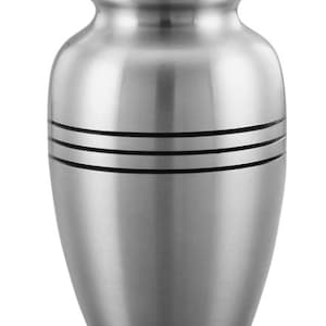 May include: A silver-colored metal cremation urn with a smooth, brushed finish. The vessel features a rounded body, a tapered neck, and a domed lid. Two distinct horizontal black bands are engraved around the upper portion of the urn's body.