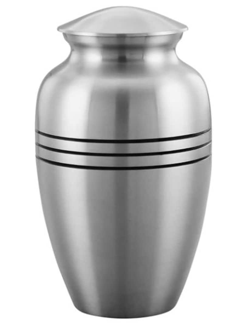 May include: A silver-colored metal cremation urn with a smooth, brushed finish. The vessel features a rounded body, a tapered neck, and a domed lid. Two distinct horizontal black bands are engraved around the upper portion of the urn's body.