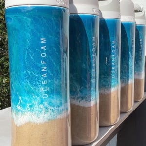 May include: A row of five cylindrical water bottles with light gray lids. Each bottle features a decorative finish resembling a beach scene with tan sand at the base, white sea foam, and a gradient of blue ocean water. The text OCEANFOAM is printed vertically on the side of each bottle.