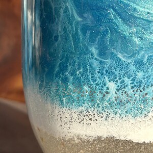 May include: A cylindrical object featuring a smooth, glossy finish with a layered design. The top section displays swirling shades of deep and light blue with a shimmering effect, transitioning into a textured white foam layer above a sandy beige base.