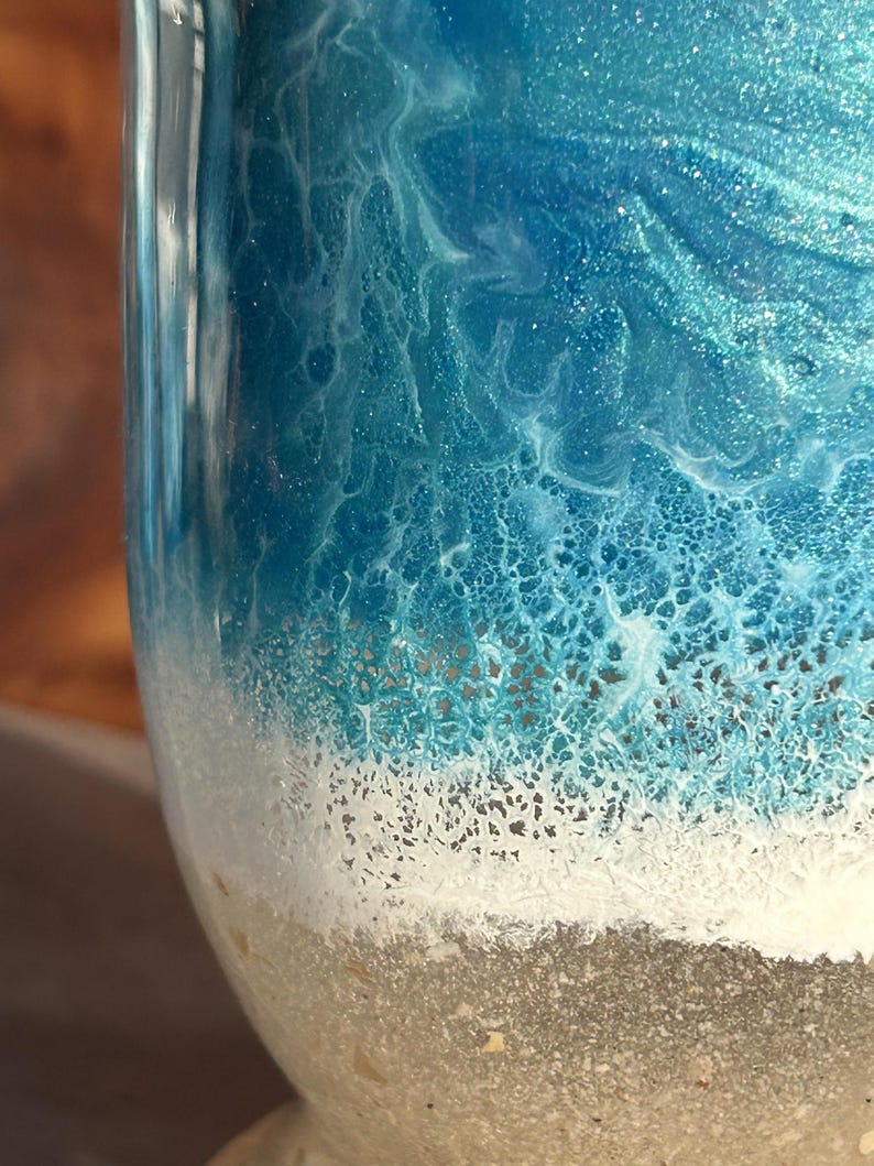May include: A cylindrical object featuring a smooth, glossy finish with a layered design. The top section displays swirling shades of deep and light blue with a shimmering effect, transitioning into a textured white foam layer above a sandy beige base.
