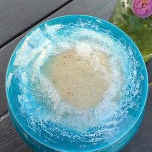 May include: A round decorative bowl featuring a glossy resin finish that mimics a coastal scene. The center is a textured beige sand color, surrounded by a vibrant turquoise blue border with white lacy foam patterns resembling ocean waves.