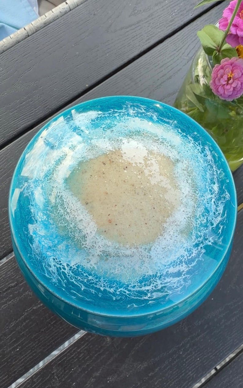 May include: A round decorative bowl featuring a glossy resin finish that mimics a coastal scene. The center is a textured beige sand color, surrounded by a vibrant turquoise blue border with white lacy foam patterns resembling ocean waves.