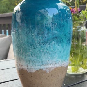 May include: A cylindrical urn with a glossy finish featuring a gradient design that transitions from a deep teal at the top to a lighter turquoise, a textured white band resembling sea foam, and a solid tan base with a speckled appearance.