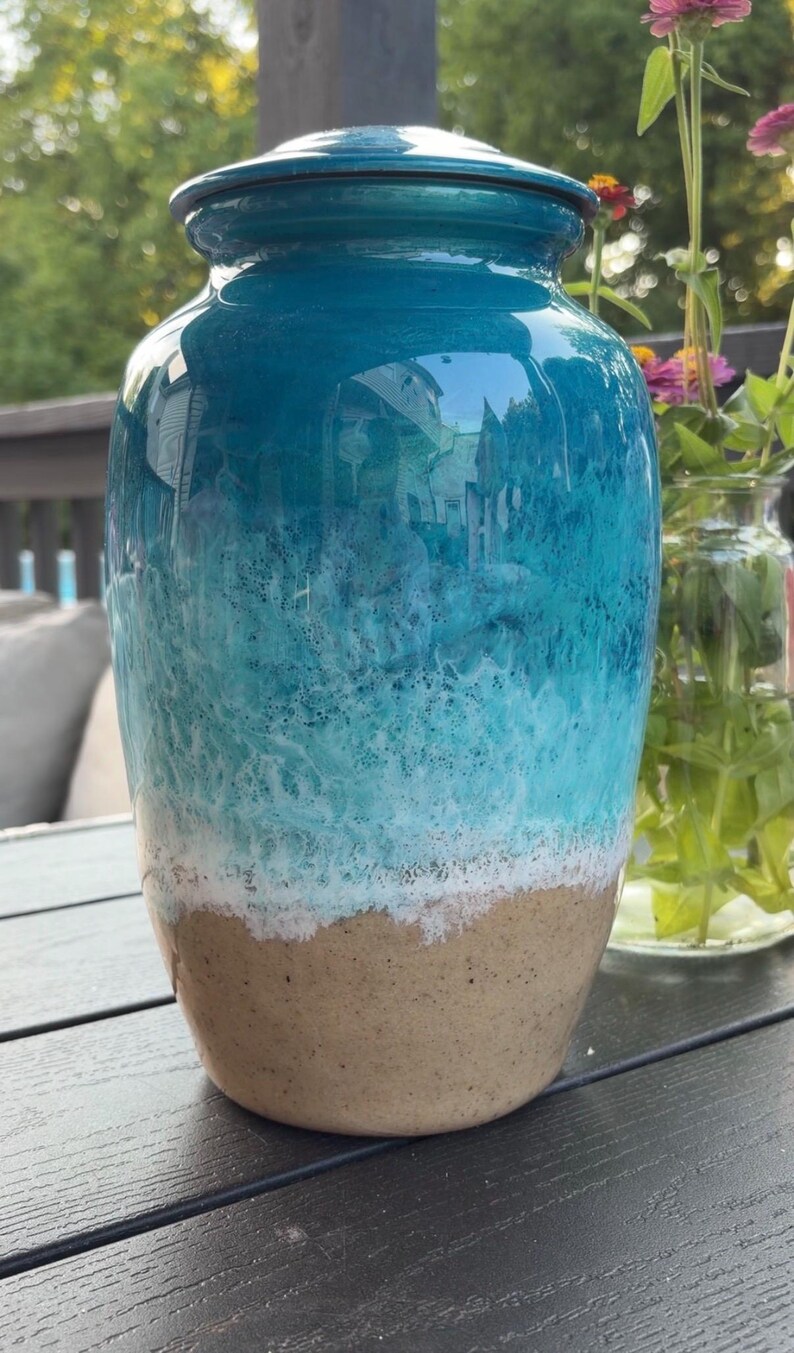 May include: A cylindrical urn with a glossy finish featuring a gradient design that transitions from a deep teal at the top to a lighter turquoise, a textured white band resembling sea foam, and a solid tan base with a speckled appearance.