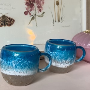 May include: Two ceramic mugs with a blue and white ocean wave design. The mugs have a sandy brown bottom and a white rim.