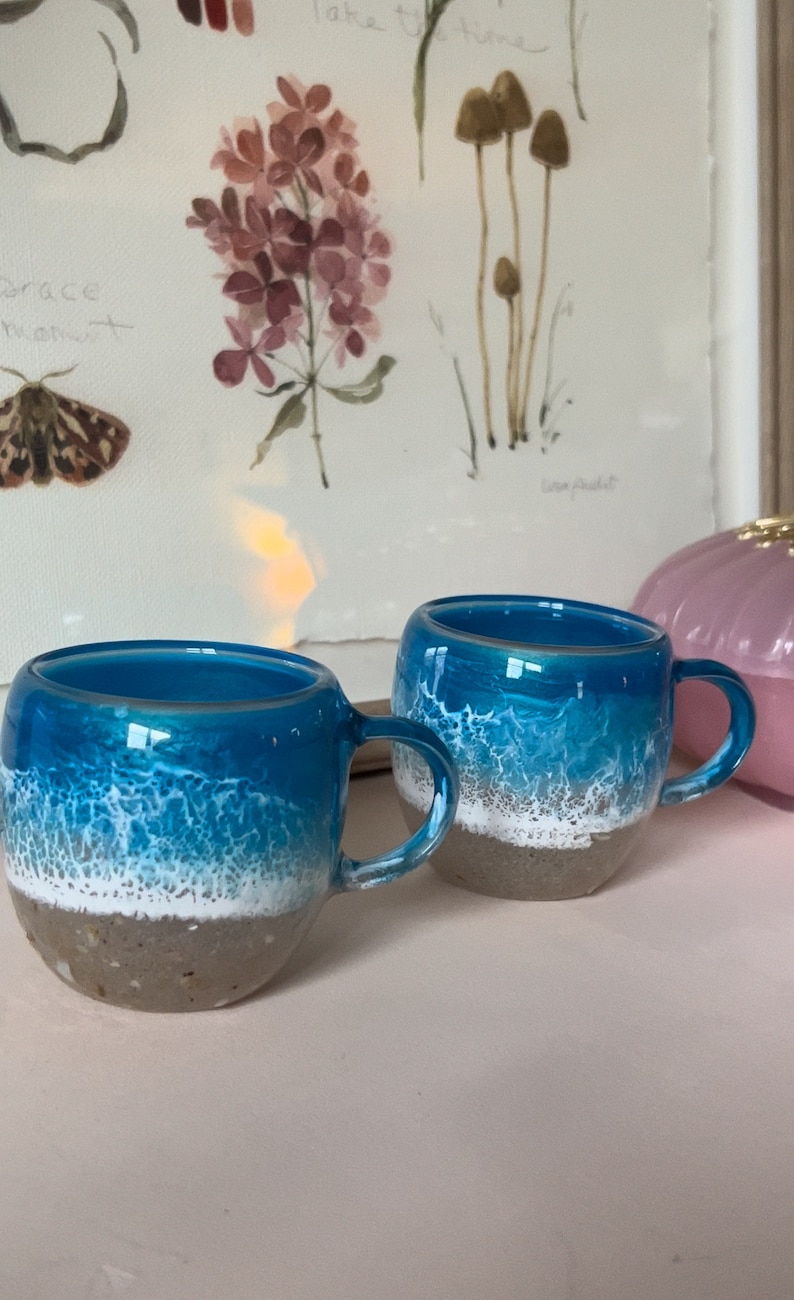 May include: Two ceramic mugs with a blue and white ocean wave design. The mugs have a sandy brown bottom and a white rim.