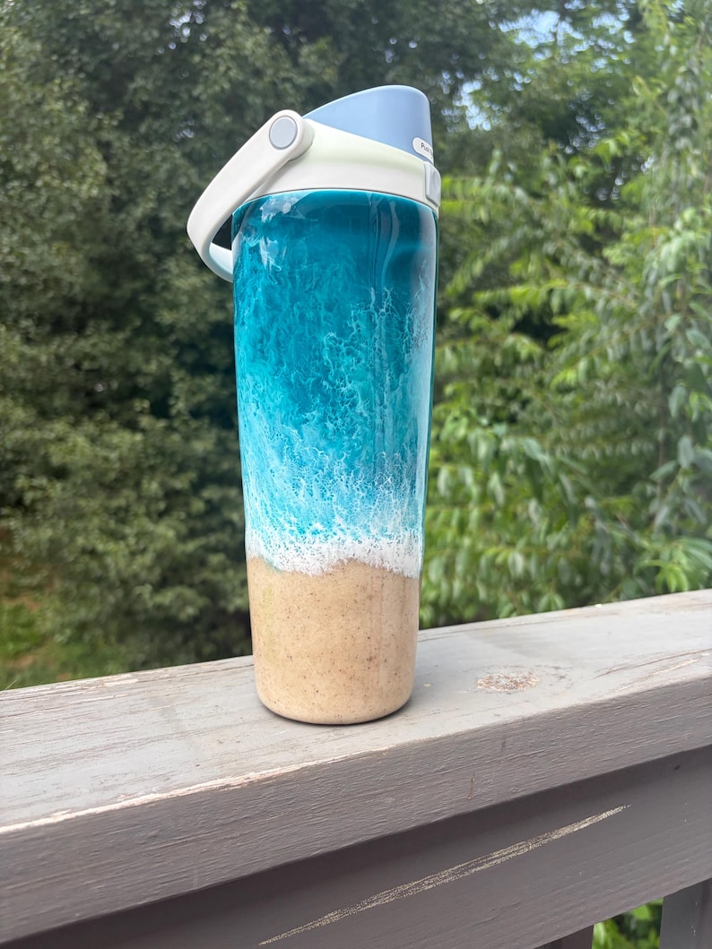 May include: A tall, insulated water bottle with a beach-themed design. The top is light blue, with a white handle and lid. The body of the bottle features a blue and white wave design that transitions to a sandy beige color at the bottom.