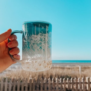 Peut inclure: Une tasse en c&eacute;ramique bleu et blanc ombr&eacute;e avec un fond sablonneux. La tasse est tenue dans une main sur fond de cl&ocirc;ture en bois et de plage.