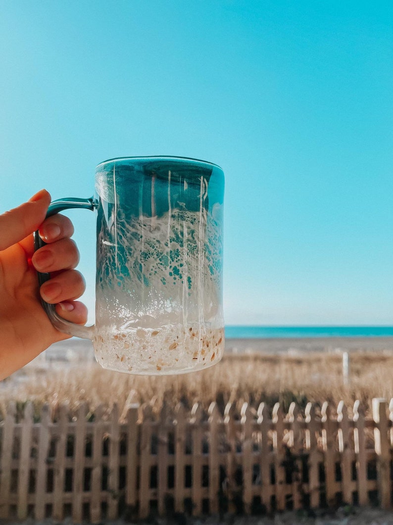Peut inclure: Une tasse en c&eacute;ramique bleu et blanc ombr&eacute;e avec un fond sablonneux. La tasse est tenue dans une main sur fond de cl&ocirc;ture en bois et de plage.