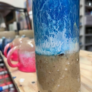 May include: A blue and white tumbler with a brown bottom. The tumbler has a white swirl design and the name "Gabby" written on it.