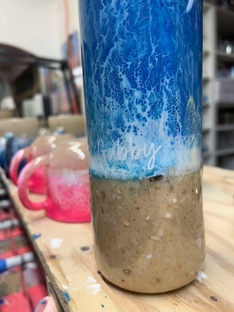 May include: A blue and white tumbler with a brown bottom. The tumbler has a white swirl design and the name "Gabby" written on it.