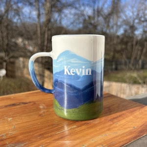 May include: White ceramic mug with a blue and green mountain scene and the name "Kevin" written in white on the front.