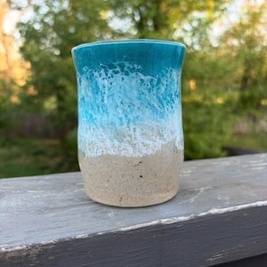 Puede incluir: Un vaso cilíndrico de cerámica con un diseño degradado que transita desde una base de color beige arena hasta una parte superior de color turquesa brillante, con un patrón blanco texturizado que simula olas rompiendo en la costa.
