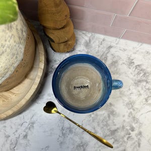 May include: A blue ceramic mug with a light brown liquid inside. The mug has the words "freckled flamingo" printed on the side. A gold heart-shaped spoon is on a white marble surface.