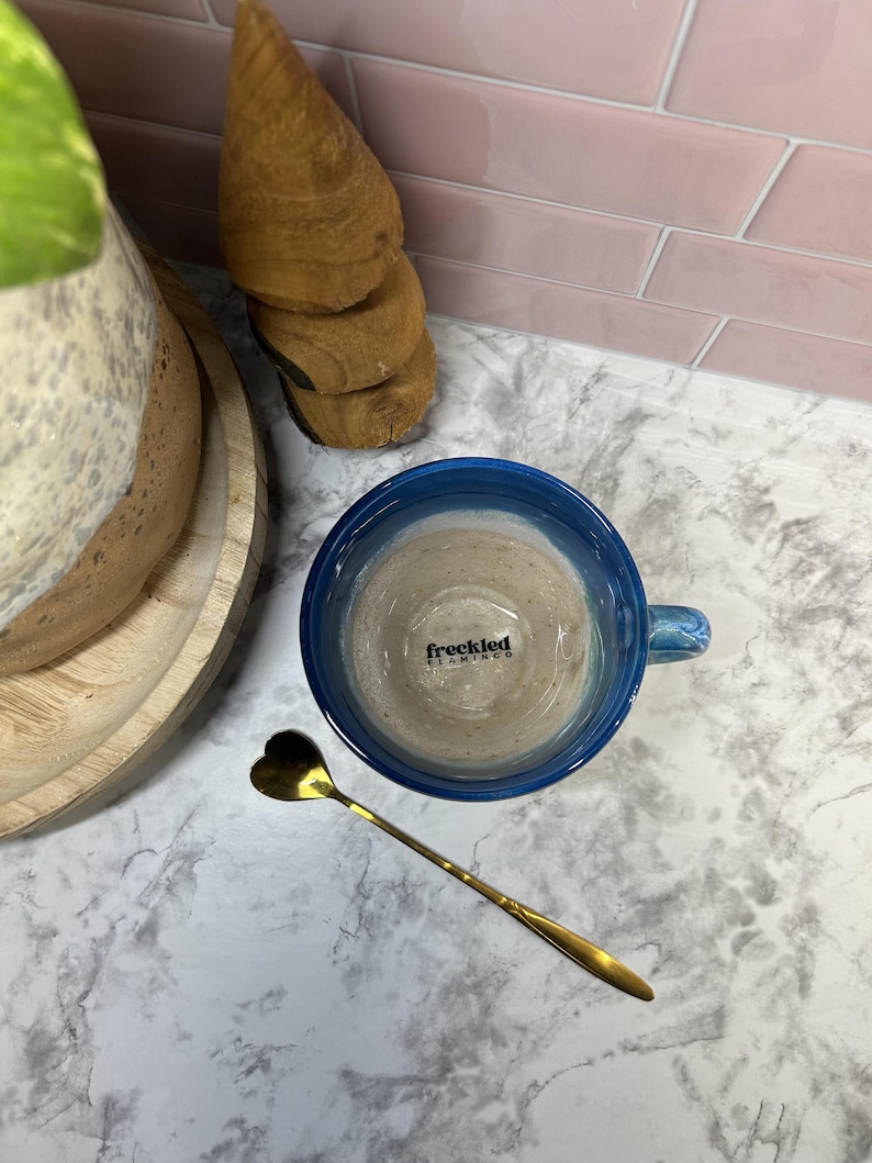 May include: A blue ceramic mug with a light brown liquid inside. The mug has the words "freckled flamingo" printed on the side. A gold heart-shaped spoon is on a white marble surface.