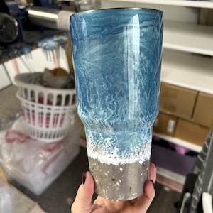 May include: A blue and white tumbler with a beach scene design. The tumbler has a silver bottom with a white rim.