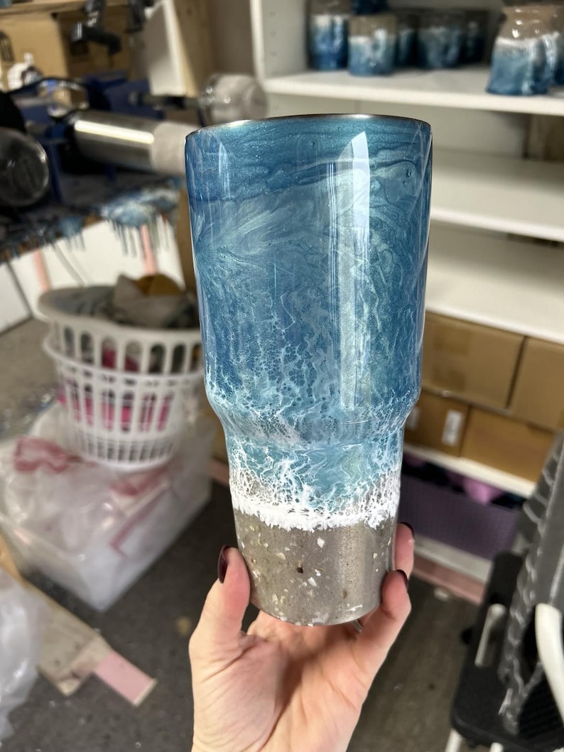 May include: A blue and white tumbler with a beach scene design. The tumbler has a silver bottom with a white rim.