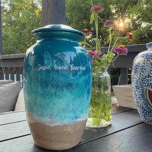 May include: A cylindrical ceramic cremation urn featuring a glossy finish with a blue and turquoise ocean wave design, a sandy beige base with a small starfish detail, and the white printed name Jason Owen Bearden on the upper section.