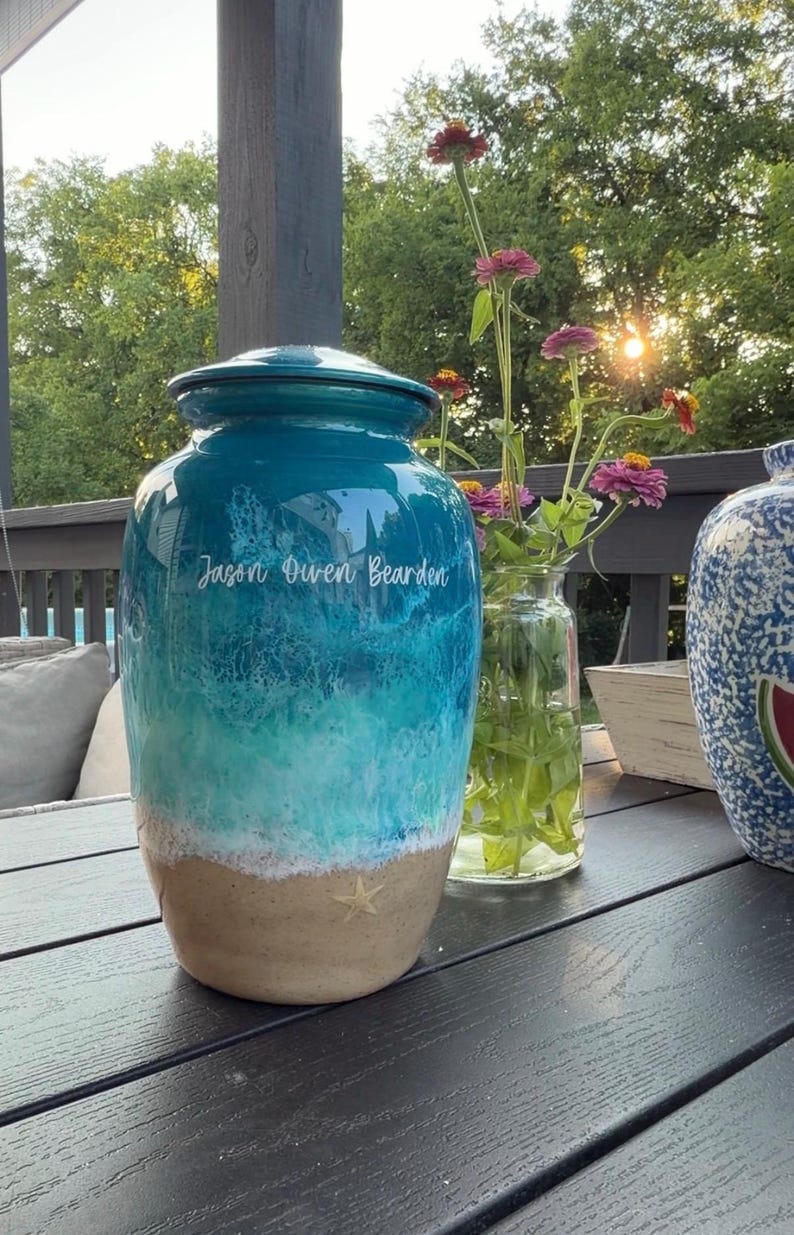 May include: A cylindrical ceramic cremation urn featuring a glossy finish with a blue and turquoise ocean wave design, a sandy beige base with a small starfish detail, and the white printed name Jason Owen Bearden on the upper section.