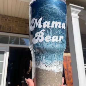 May include: A blue and white ocean-themed tumbler with the text "Mama Bear" in white lettering. The tumbler has a clear lid and a sandy bottom.