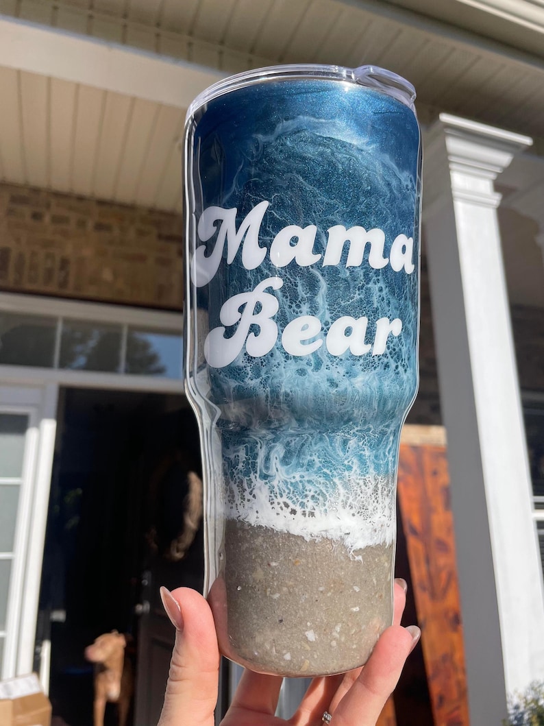 May include: A blue and white ocean-themed tumbler with the text "Mama Bear" in white lettering. The tumbler has a clear lid and a sandy bottom.