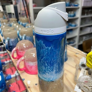 May include: A white and blue water-themed insulated water bottle with a gray lid and the word "owala" printed on the side. The bottle has a white and blue design that resembles waves and a sandy beach at the bottom.