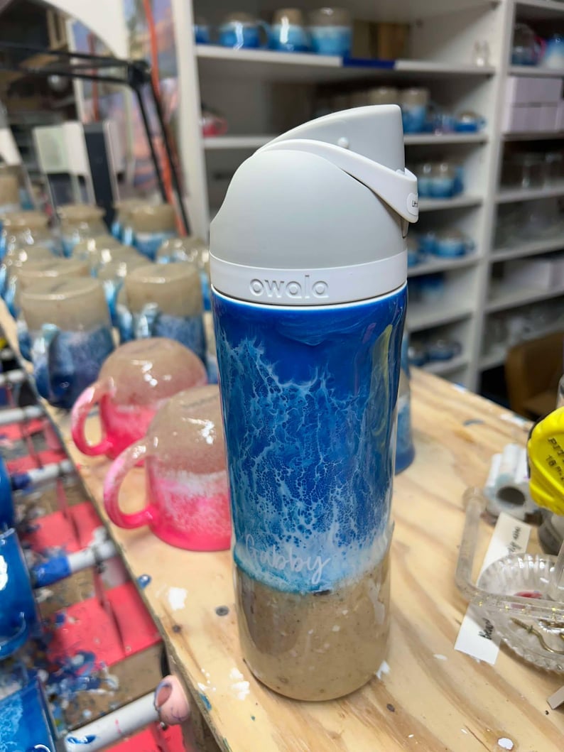 May include: A white and blue water-themed insulated water bottle with a gray lid and the word "owala" printed on the side. The bottle has a white and blue design that resembles waves and a sandy beach at the bottom.