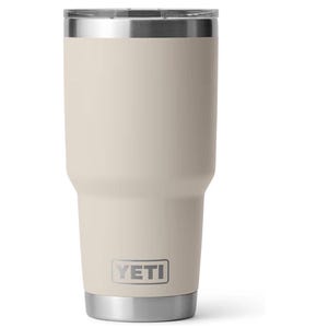 May include: A beige stainless steel tumbler with a tapered body and a clear lid. The exterior features a matte finish with the YETI logo printed in a rectangular frame near the base. The top rim and the base are made of polished silver metal.