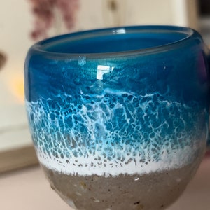 May include: A blue and white resin cup with a beach scene design. The cup has a white foamy wave on top of a blue ocean, and a brown sandy bottom.