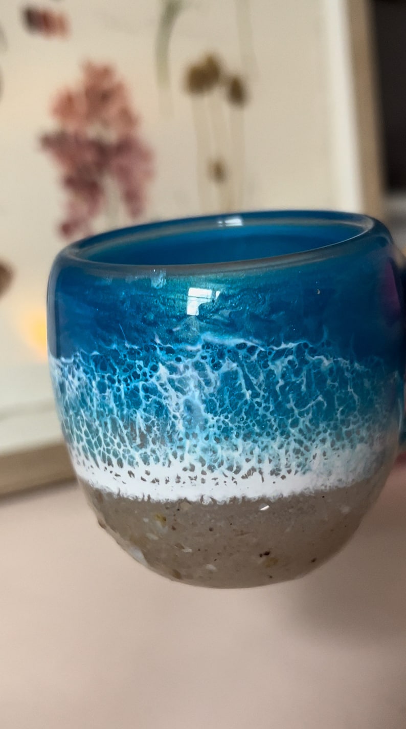 May include: A blue and white resin cup with a beach scene design. The cup has a white foamy wave on top of a blue ocean, and a brown sandy bottom.