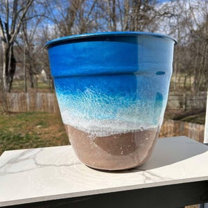 Può includere: Un vaso da fiori in ceramica blu, bianco e marrone con un design a tema spiaggia. Il vaso è appoggiato su una superficie di marmo bianco.